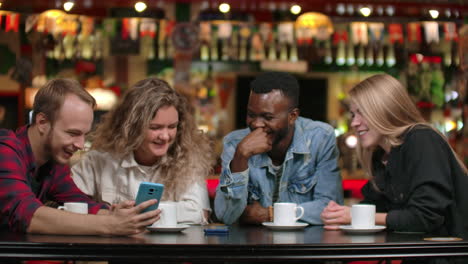 Afroamerikanischer-Mann-Und-Mädchen-Schauen-Auf-Ein-Smartphone-Und-Unterhalten-Sich,-Während-Sie-In-Einem-Café-Sitzen-Und-Kaffee-Trinken,-Mit-Freunden-Und-Einer-Gruppe-Von-Leuten-In-Einem-Restaurant.